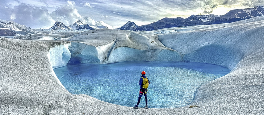 A hiker stands before a vivid blue glacial pool set within sweeping ice fields framed by rugged Patagonian peaks.