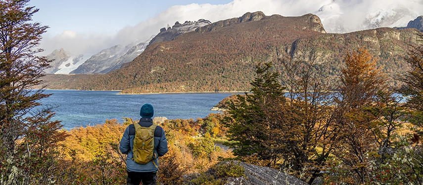 A traveller looks across a lake towards forested slopes and distant snowy mountains glowing under soft Patagonian light.