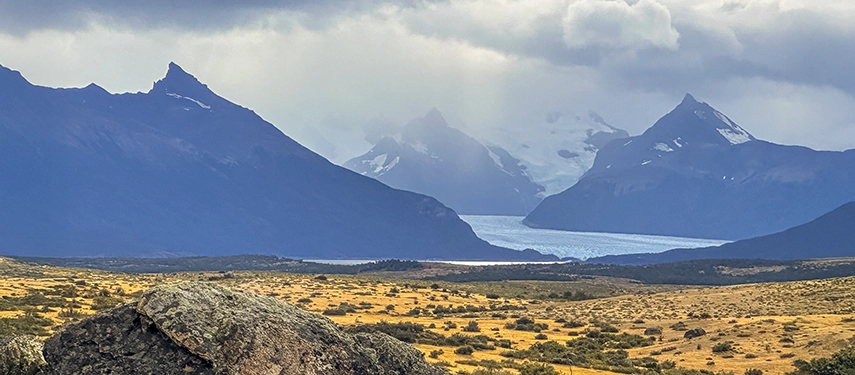 Golden plains stretch towards a distant glacial lake framed by dark mountain silhouettes beneath a brooding sky.