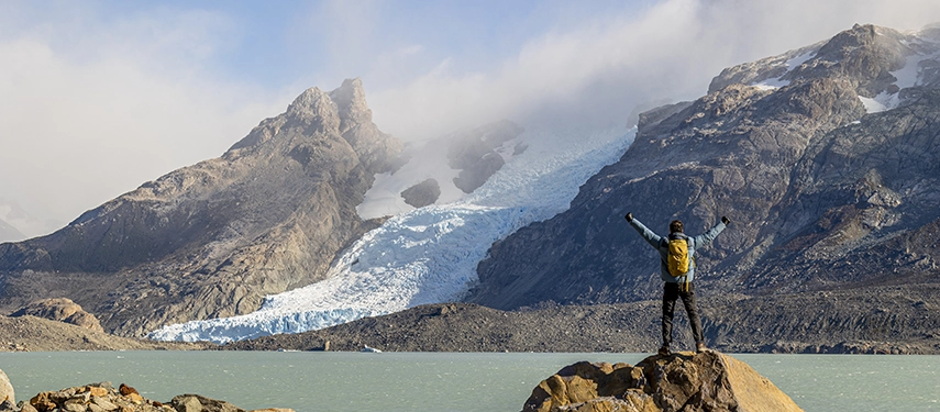 A traveller stands on a rock before a rugged glacier, arms raised towards the dramatic peaks and icy landscape beyond.