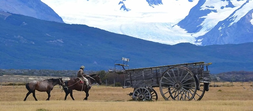 Gaucho on horseback leading a second horse past an old wooden cart with snowy peaks beyond.