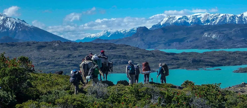 Group of hikers and horse riders travelling along a ridge with turquoise lakes and snowy mountains behind.