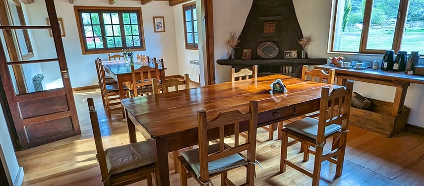 Rustic dining room with wooden tables and chairs, bright windows, and a traditional fireplace.