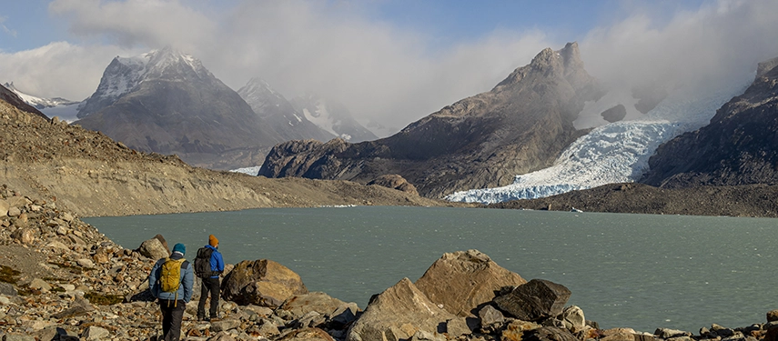 Two hikers follow a rocky lakeshore beneath towering peaks and a sweeping glacier under shifting Patagonian cloud.