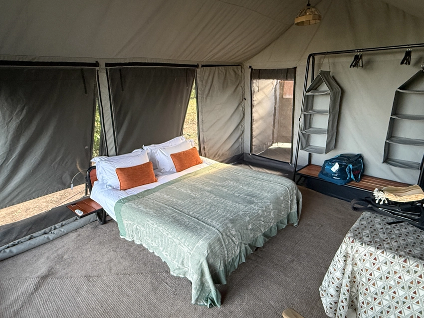 Inside a classic safari tent, a generously dressed bed and simple furnishings create a calm, grounded retreat surrounded by nature.