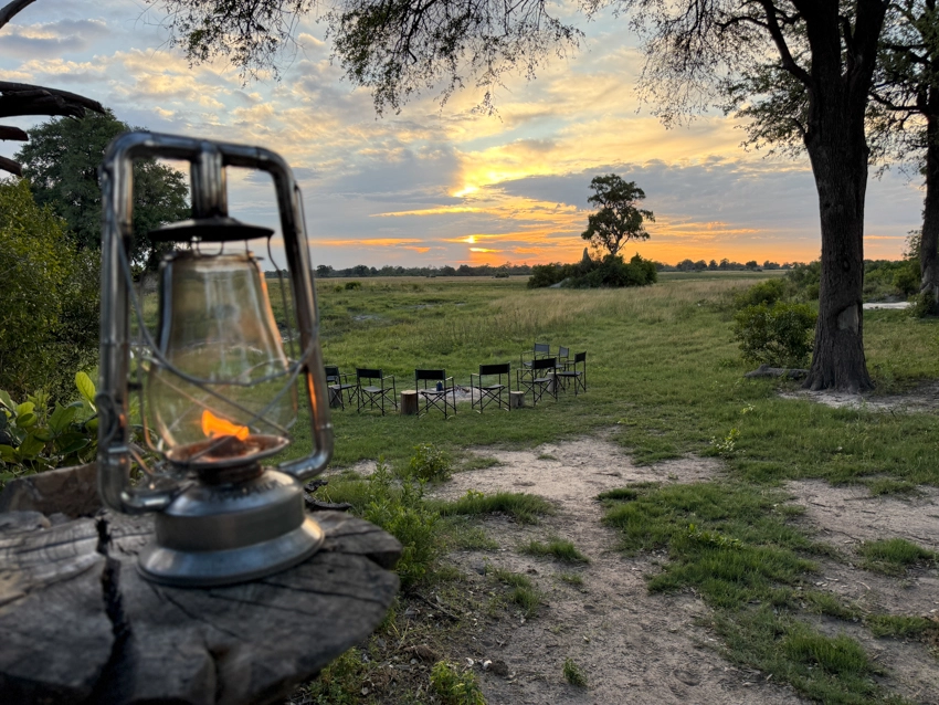 A lantern glows at dusk beside a circle of safari chairs, set for evening storytelling as the sun sets over the open floodplains.