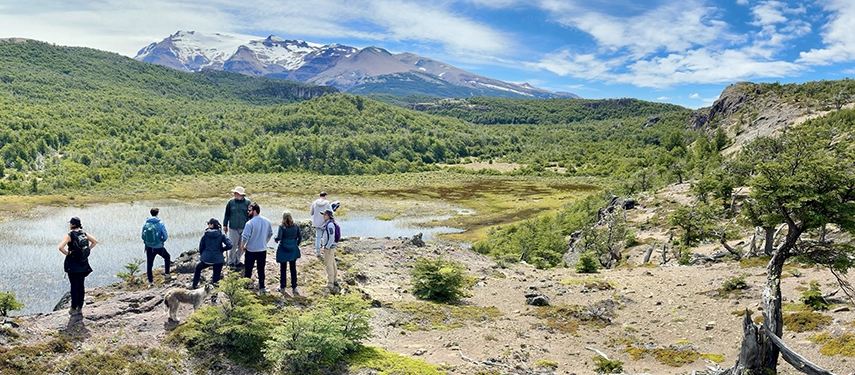 Group of hikers and a dog overlooking a small mountain lake with snow-capped peaks in the distance.