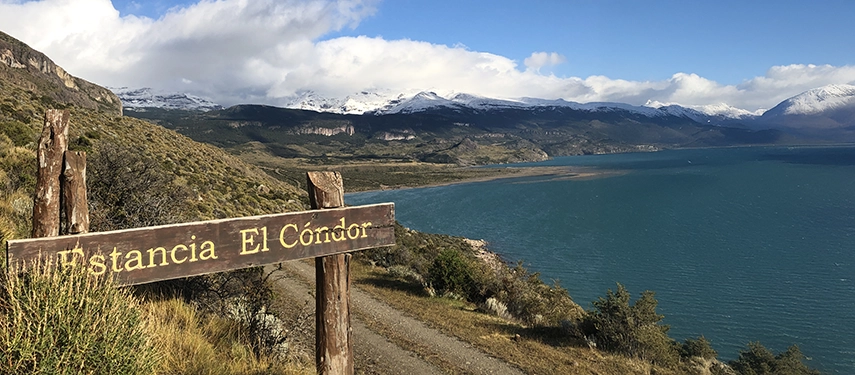 Wooden sign for Estancia El Cóndor with Lago San Martín and snow-covered mountains in the background.
