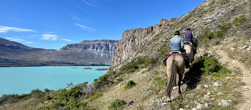 Two riders on horseback following a cliffside trail above the turquoise waters of Lago San Martín.
