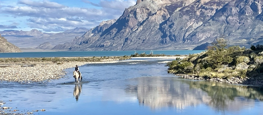 Horse and rider crossing a shallow river with dramatic Patagonian mountains rising in the background.