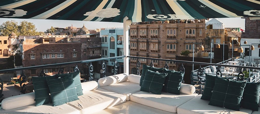 Rooftop lounge with vibrant umbrella shade and views of historic Jodhpur buildings.