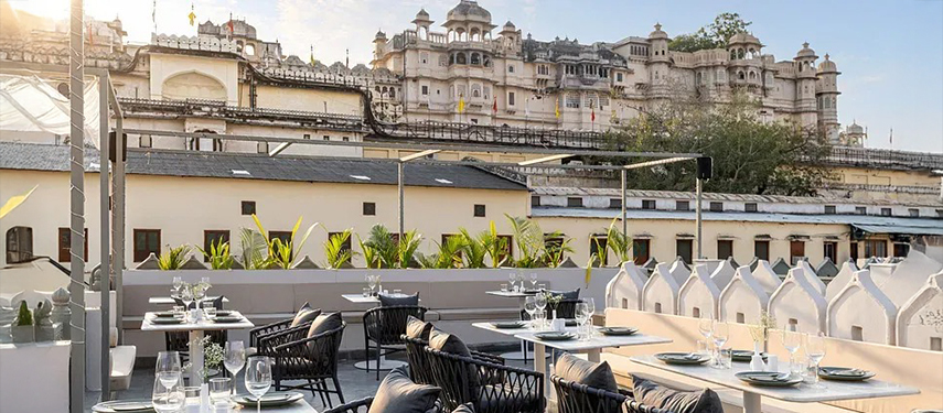 Daytime rooftop dining area with black wicker seating and palace backdrop at Jhilwara Haveli