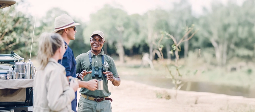 Guide chatting with travellers beside a safari vehicle during a morning stop in the bush.
