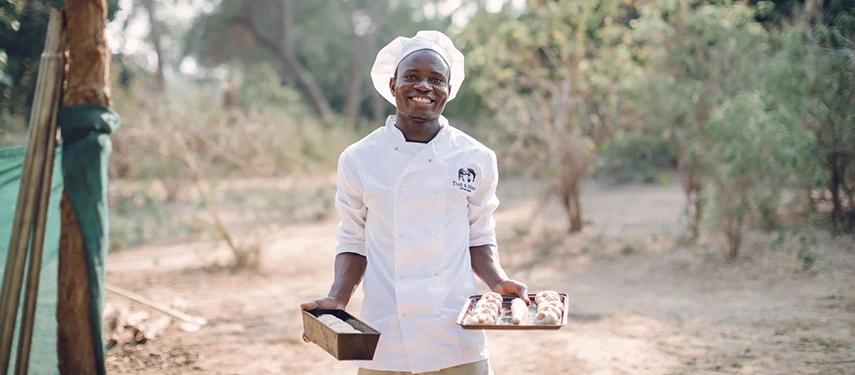 Smiling camp chef holding trays of freshly baked bread and pastries at Kutali Camp.