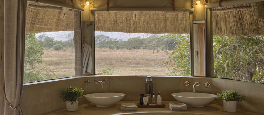 Twin vessel basins sit beneath a wide, reed-framed window in a Lion Camp suite bathroom, looking out across open South Luangwa grasslands — the wilderness framed as naturally as any artwork.