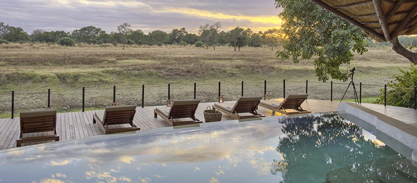 Lion Camp's infinity pool and timber sun deck face out across the South Luangwa floodplains at dusk, sunloungers reflected in the still water as the sky turns gold and cloud-streaked above the treeline.