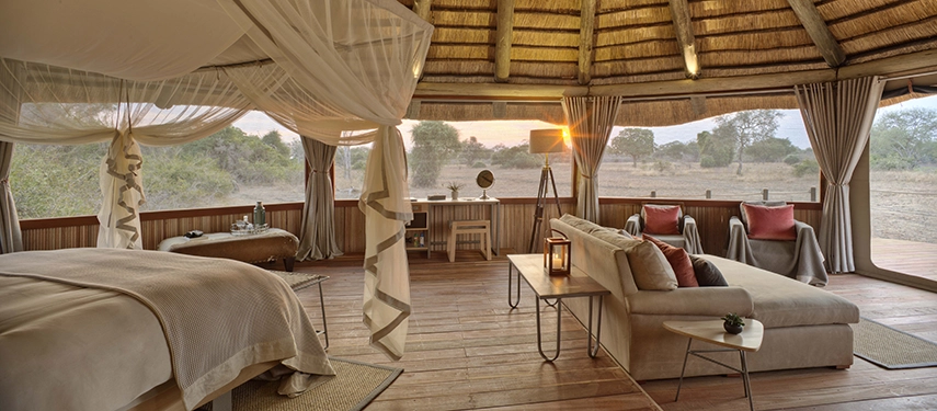 The open, circular interior of a Lion Camp deluxe suite at dusk — a four-poster king bed draped in white muslin, a generous lounge area, and canvas walls rolled back to reveal an unbroken view across the South Luangwa plains.