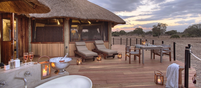 A Lion Camp deluxe suite deck glows with candlelight at dusk — a freestanding bathtub, champagne on ice, sunloungers, and a private dining table set against an open horizon of South Luangwa bushveld.