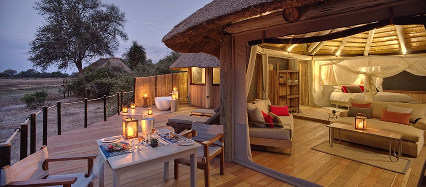 At dusk, a private dinner table is set on a Lion Camp deluxe suite deck, lanterns glowing beside a freestanding bathtub and sunloungers, the warm interior of the suite and the open bushveld beyond visible in the same frame.
