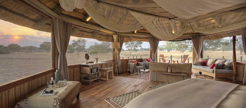 Looking out from inside a Lion Camp deluxe suite at sunset, canvas walls open fully to the South Luangwa grasslands, a king bed in the foreground and lounge chairs on the private deck catching the last of the day's light.