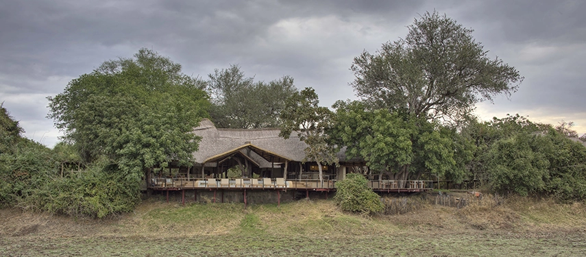 Lion Camp's thatched main lodge sits elevated above the South Luangwa floodplain on raised timber supports, its open-sided deck and lounge area nestled among mature trees beneath a heavy, cloud-filled sky.