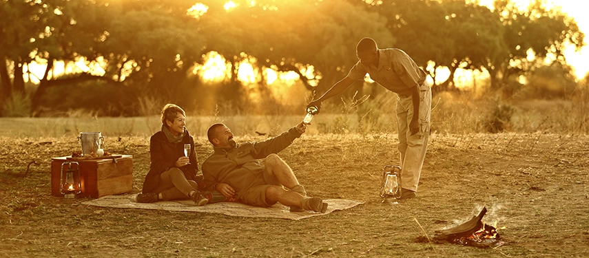 A couple recline on a blanket in the open South Luangwa bush at golden hour, a staff member pouring champagne beside a small fire and a wooden drinks table as the sun sets through the trees behind them.