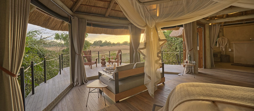 A Lion Camp luxury suite interior opens towards the South Luangwa bush at golden hour, sheer curtains drifting at the open canvas walls, a private deck with chairs visible beyond the foot of the king-sized bed.