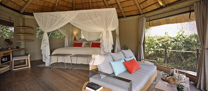 Inside a thatched Lion Camp luxury suite, a four-poster bed with white muslin canopy is paired with a generous sofa dressed in colourful cushions, the whole space enclosed by a circular thatched roof and bush views on every side.