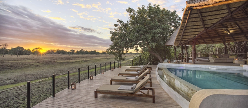 Lion Camp's curved swimming pool and long timber sun deck face west across the South Luangwa floodplain at sunset, sunloungers lined along the water's edge and the thatched main lodge canopy framing the scene to the right.