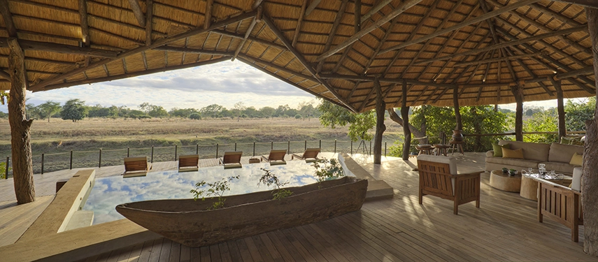 Beneath a soaring thatched roof, Lion Camp's main lodge lounge and pool area open onto a wide timber deck facing the South Luangwa plains, the curved pool reflecting a cloud-streaked sky and the bush stretching to the horizon beyond.