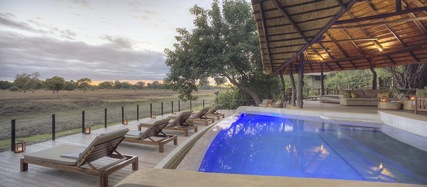 Lion Camp's illuminated pool glows blue at dusk, sunloungers and lanterns arranged along the timber deck while the South Luangwa floodplain stretches away beneath a fading pink and amber sky, the thatched lodge canopy overhead.