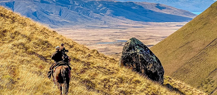 A rider ascends a steep, sunlit hillside overlooking vast plains and distant ridges of the Patagonian steppe.