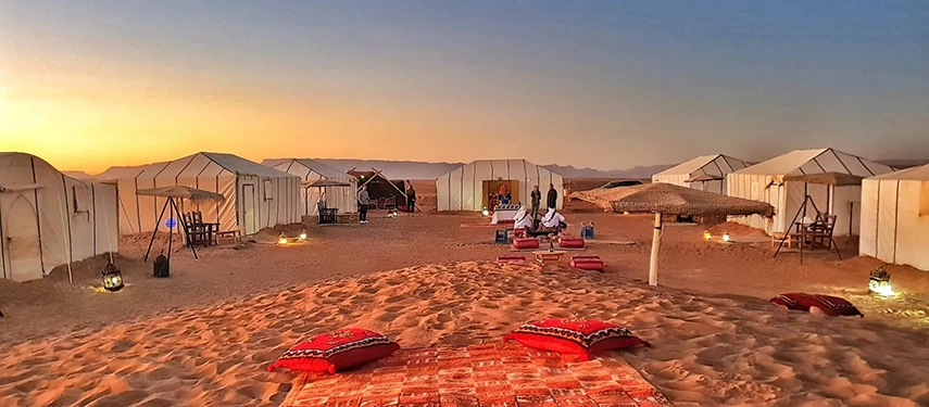 White canvas tents illuminated at dusk with lanterns and seating areas in the sandy desert of Merzouga.
