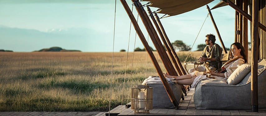 Guests relax on cushioned loungers on a private tented deck, gazing out across golden grasslands in the soft light of late afternoon.