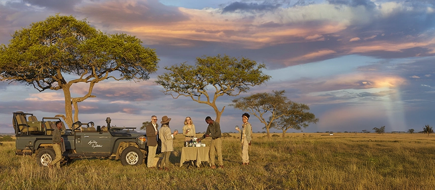 A safari vehicle is parked beside a pop-up sundowner table on the open plains, framed by acacia trees and a dramatic sunset sky.
