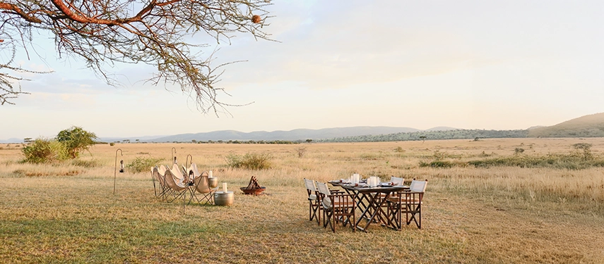 An elegant outdoor dining table is set on the savannah, surrounded by open grassland and distant hills under a calm evening sky.
