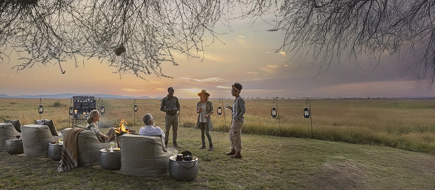 Guests gather around a firepit and al fresco bar at dusk, lanterns glowing as the Western Serengeti stretches into the horizon.