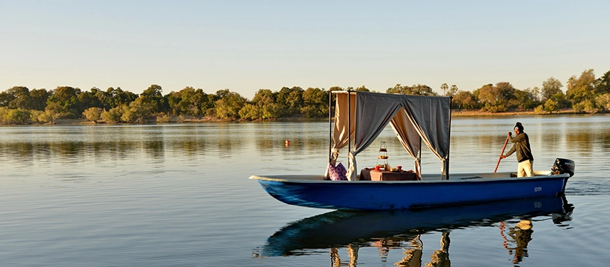 A private boat drifts along the Zambezi River as guests enjoy an elegant high tea experience surrounded by calm waters and riverine forest.