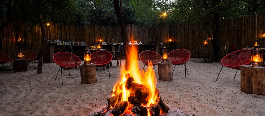 The boma at Mukwa River Lodge glows at night, with a central fire pit, lantern light, and relaxed seating beneath the trees.