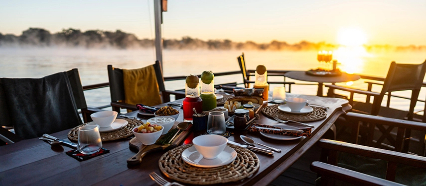 Breakfast is served on the Zambezi River during a river safari, with beautifully set tables overlooking misty water at sunrise.