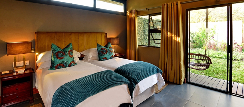 Twin beds in a Mukwa River Lodge cottage are dressed in crisp linens and rich textures, opening onto a garden terrace with a hanging chair.