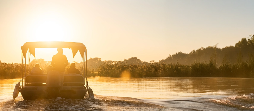 A small boat cruises the Zambezi River at golden hour, capturing the serene atmosphere of a river safari near Mukwa River Lodge.