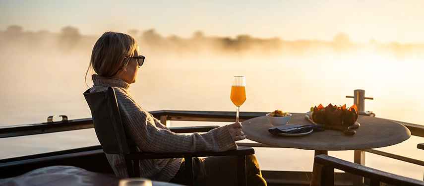 A guest relaxes with a drink on a river safari at sunrise, mist lifting from the Zambezi River during a tranquil morning experience.