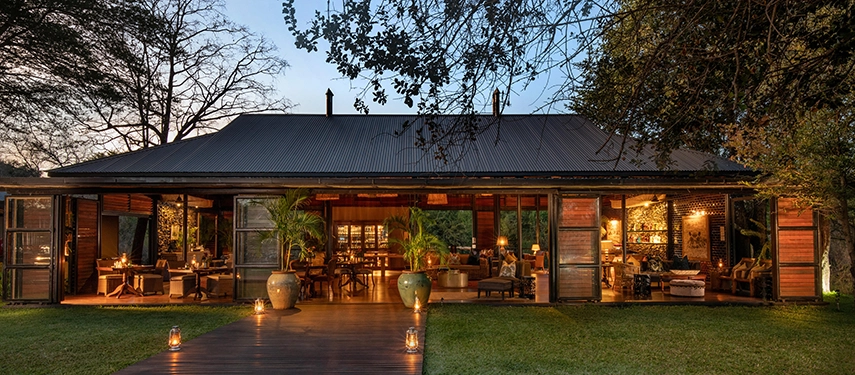 Warm light spills from the open-fronted lodge at Mukwa River Lodge, highlighting its elegant design and tranquil garden setting after sunset.