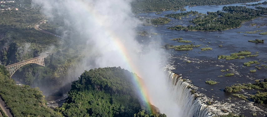 A dramatic aerial perspective captures Victoria Falls with rising spray and a rainbow arcing above the Zambezi River, close to Mukwa River Lodge.