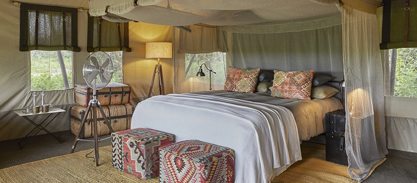 The tented bedroom at Nyasi Tented Camp features soft linens, patterned textiles, and large mesh windows opening to the Serengeti landscape.