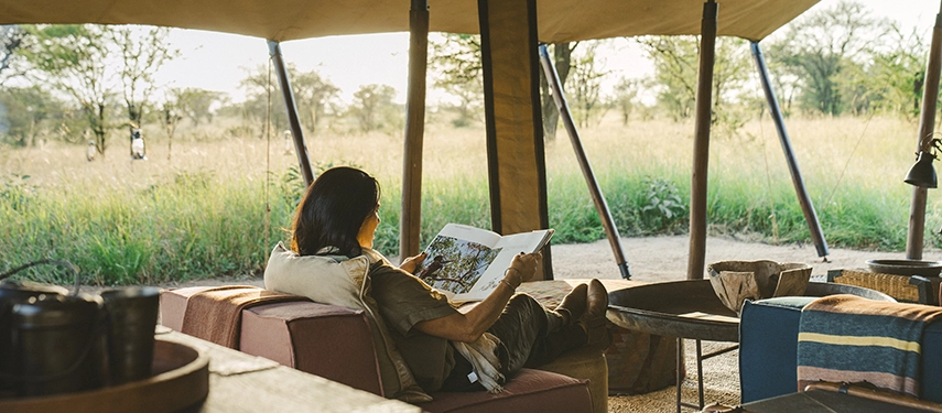 A quiet moment in the Nyasi Tented Camp lounge shows a guest reading while seated beneath canvas with views across the surrounding grasslands.