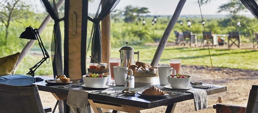 Breakfast is served beneath canvas at Nyasi Tented Camp, with fresh dishes laid out on a table overlooking the grassy Serengeti landscape.