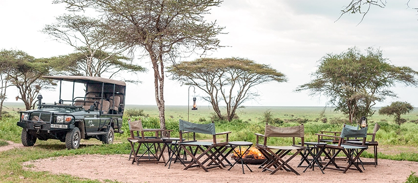 Safari chairs circle a glowing campfire beside a parked game drive vehicle, capturing the timeless evening ritual at Nyasi Tented Camp.