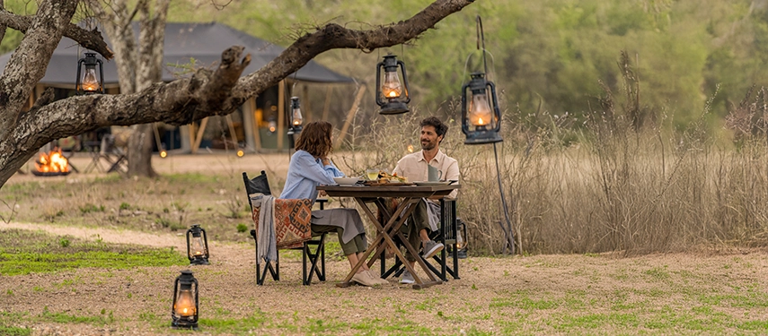 An intimate outdoor dinner setting at Nyasi Tented Camp is illuminated by lanterns, creating a warm evening atmosphere beneath the trees.